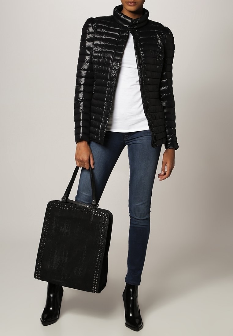 Femme portant une doudoune noire, chemise blanche, jean skinny bleu, bottines noires, tenant un grand sac fourre-tout noir clouté.