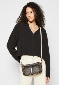 Czarna krótka bluzka z zapięciem na guziki, zestawiona z brązową torebką na ramię w wzory, z złotymi elementami i łańcuszkowym paskiem. Białe spodnie.