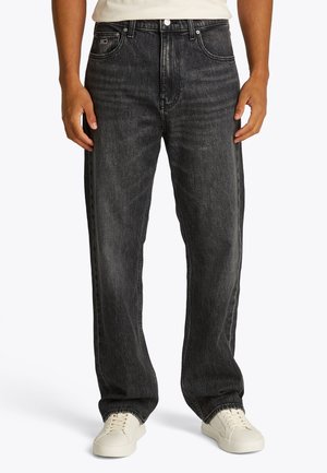 Άνδρας που φοράει φαρδιά μαύρα τζιν και λευκά αθλητικά παπούτσια, στέκεται με τα χέρια χαλαρά στα πλάγια σε λευκό φόντο.