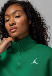 Zielona bluza na zamek z wysokim kołnierzem, z białym logo Jumpman na piersi i teksturowaną tkaniną. Zbliżenie na linię szyi.