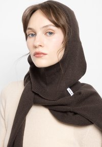 Brązowy dzianinowy szalik z kapturem, o prążkowanej strukturze i widoczną metką z logo. Nosi się go na kremowym swetrze.