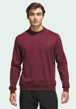 Giovane uomo con felpa Adidas bordeaux a maniche lunghe e pantaloni neri, in piedi davanti a uno sfondo chiaro e semplice.