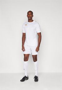 Jersey atlético blanco y pantalones cortos con acentos dorados. Cuenta con un escote redondo, mangas cortas y botas de fútbol negras.