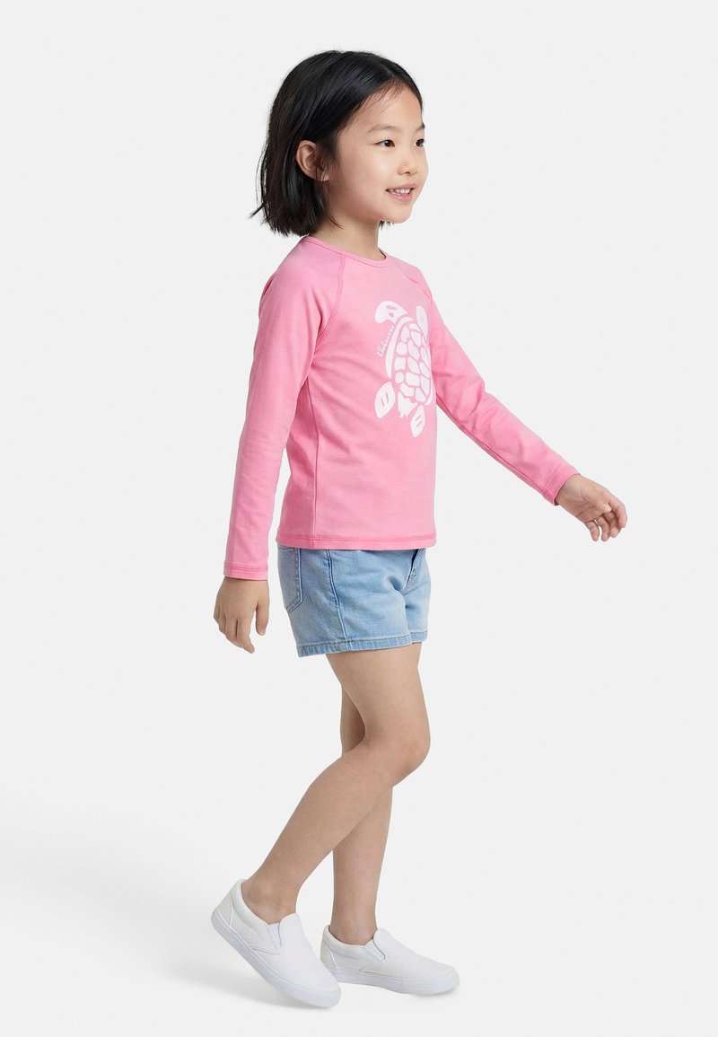 Κορίτσι που χαμογελά περπατώντας, φορώντας ροζ μπλούζα με μακριά μανίκια και σχέδιο χελώνας, ανοιχτό μπλε σορτς και άσπρα παπούτσια slip-on, σε λευκό φόντο.