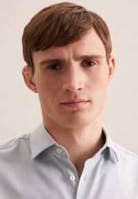 Camicia button-down azzurro chiaro con un motivo testurizzato. Presenta un colletto aperto ed è indossata da una persona con capelli corti castani, su uno sfondo neutro.
