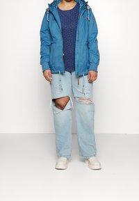 Veste légère bleue avec capuche, poignets élastiques et cordons de serrage. Chemise bleu foncé à pois blancs. Jean délavé bleu clair et chaussures beiges.