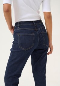 Jeans en denim bleu foncé à coupe Slim, dotés de poches arrière et d'accents de couture visibles. Texture légèrement délavée sur le tissu.
