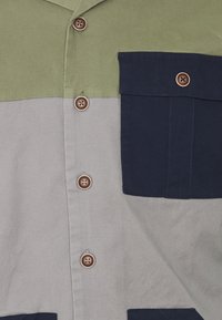 Camicia button-up con blocchi di colore verde oliva, grigio e blu navy. Quattro bottoni con finitura testurizzata, una tasca a toppa in blu navy.