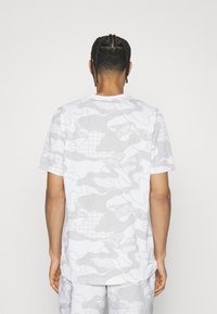 Mężczyzna z warkoczami, ubrany w koszulę z krótkim rękawem w biało-szary kamuflaż oraz pasujące szorty, stojący tyłem na jednolitym tle.