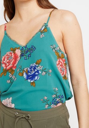 Kobieta ubrana w turkusową, bezrękawnikową bluzkę w kwiaty różowego i niebieskiego koloru oraz oliwkowozielone spodnie z wiązanym paskiem w talii.