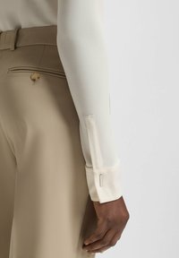 Beige tailored trousers with a back pocket and button detail, paired with a light cream long-sleeve blouse featuring buttoned cuffs.