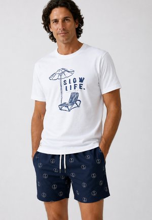 Hombre con camiseta blanca con el texto "Slow Life" y diseño de silla de playa, pantalones de baño azul marino con patrones blancos de anclas, manos en los bolsillos.