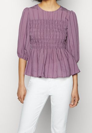 Femme portant un chemisier violet smocké avec des manches bouffantes trois-quarts et un pantalon blanc, debout devant un fond uni.