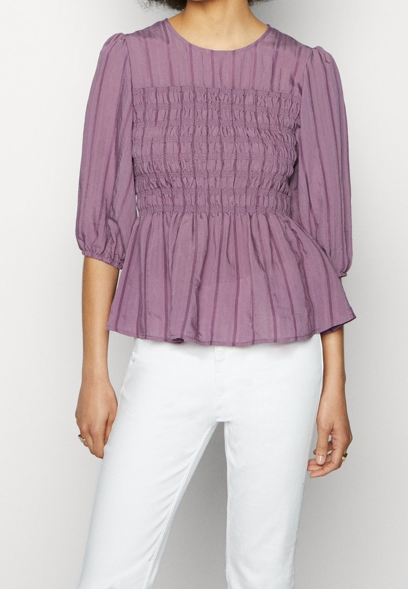 Femme portant un chemisier violet smocké avec des manches bouffantes trois-quarts et un pantalon blanc, debout devant un fond uni.