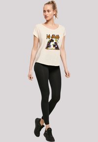 Cremefarbenes, kurzärmliges T-Shirt mit einem bunten grafischen Design, das eine Pyramide und Text zeigt, kombiniert mit schwarzen Leggings und Schuhen.