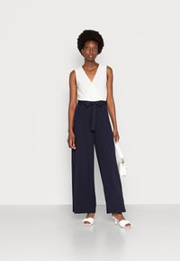 Macacão azul-marinho com corpete envolvente branco, pernas largas e um laço na cintura. Apresenta uma textura suave e um design minimalista.