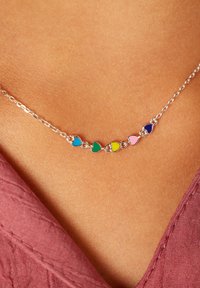 Zilveren ketting met zeven kleurrijke hartvormige bedels in blauw, groen, geel, roze en paars. Gladde textuur, minimaal ontwerp.