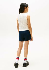 Top sans manches crème en tricot avec un motif texturé, associé à un short marine. Le modèle porte des chaussettes blanches avec des rayures rouges et bleues et des chaussures noires.
