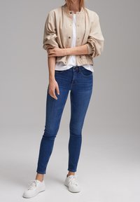 Beige bomber bunda s elastickými manžetami, nošená přes bílou košili, spárovaná s modrými skinny džíny a bílými teniskami.