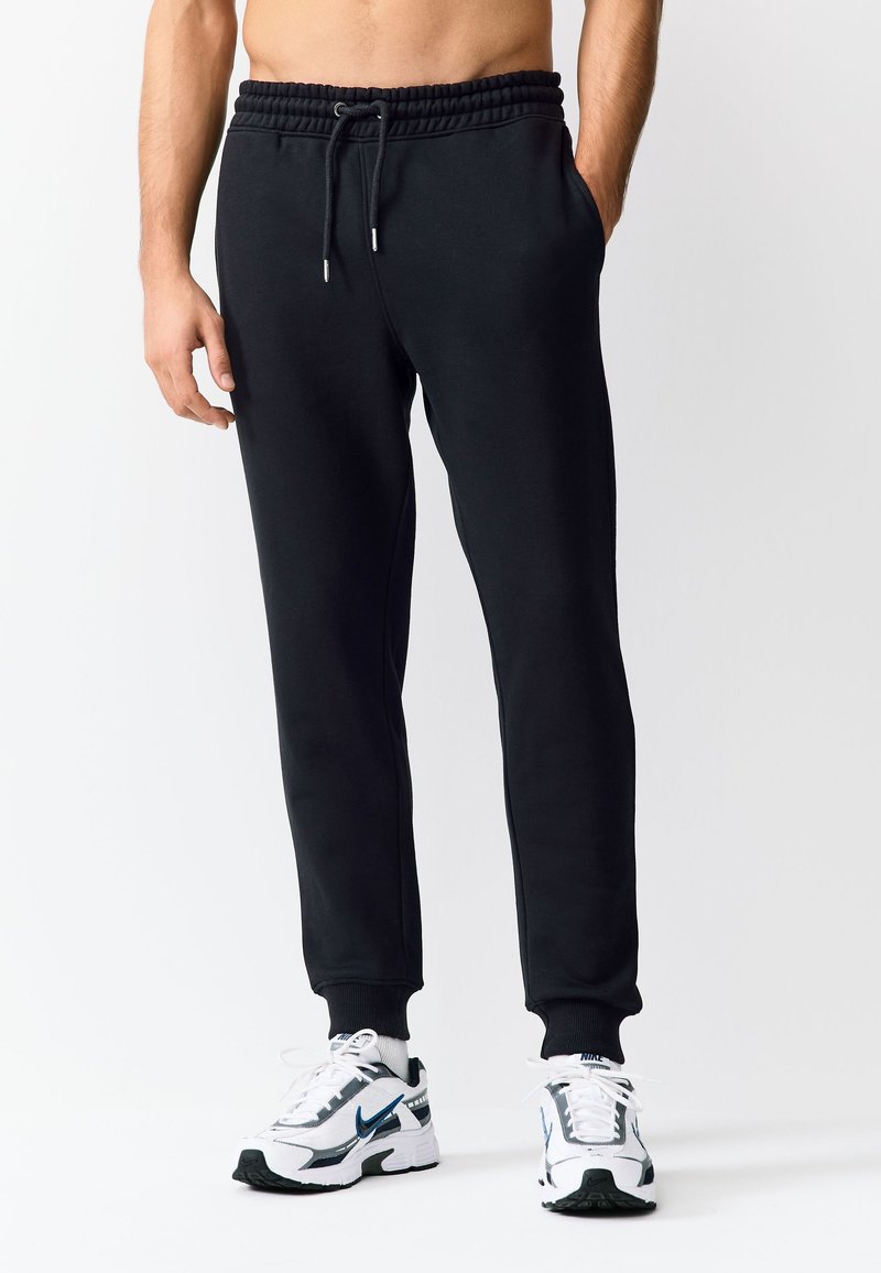 Homme portant un pantalon de jogging noir et des baskets blanches Nike, debout devant un fond clair uni, les mains dans les poches.