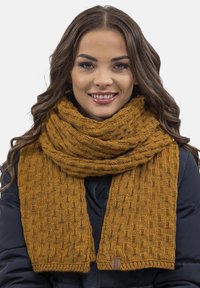 Bufanda de color mostaza tejida, caracterizada por un patrón texturizado, lana gruesa y una etiqueta. Llevada de manera suelta alrededor del cuello.