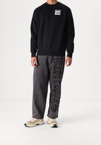Felpa nera con collo a giro e patch logo bianco. Jeans grigio scuro con grafica testuale nera grande su una gamba. Sneaker beige con dettagli neri.