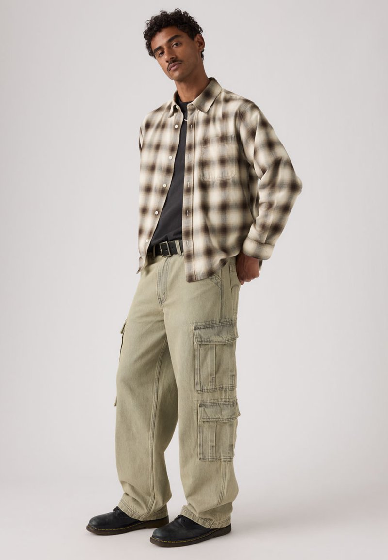 Gefüttertes Hemd mit Kragen in beige-braunem Karomuster, kombiniert mit olivgrünen Cargo-Hosen mit mehreren Taschen und schwarzen Schuhen.