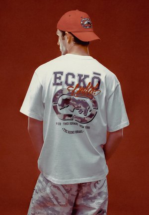 White short-sleeve t-shirt with graphic print on back, featuring purple and orange lettering, paired with camo-print shorts and a red cap.