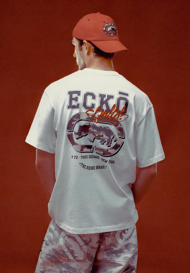 White short-sleeve t-shirt with graphic print on back, featuring purple and orange lettering, paired with camo-print shorts and a red cap.