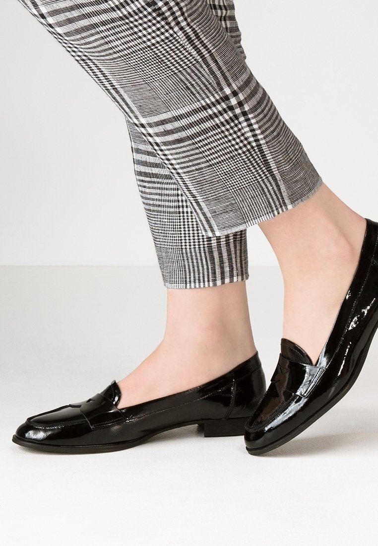 Pieds portant des mocassins en cuir verni noir associés à un pantalon court à carreaux noir et blanc sur un fond uni.
