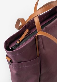 Leder-Tote-Bag in tiefem lila, mit braunen Lederträgern, einem Reißverschluss oben und Außentaschen, mit glatter Textur und Nähdetails.