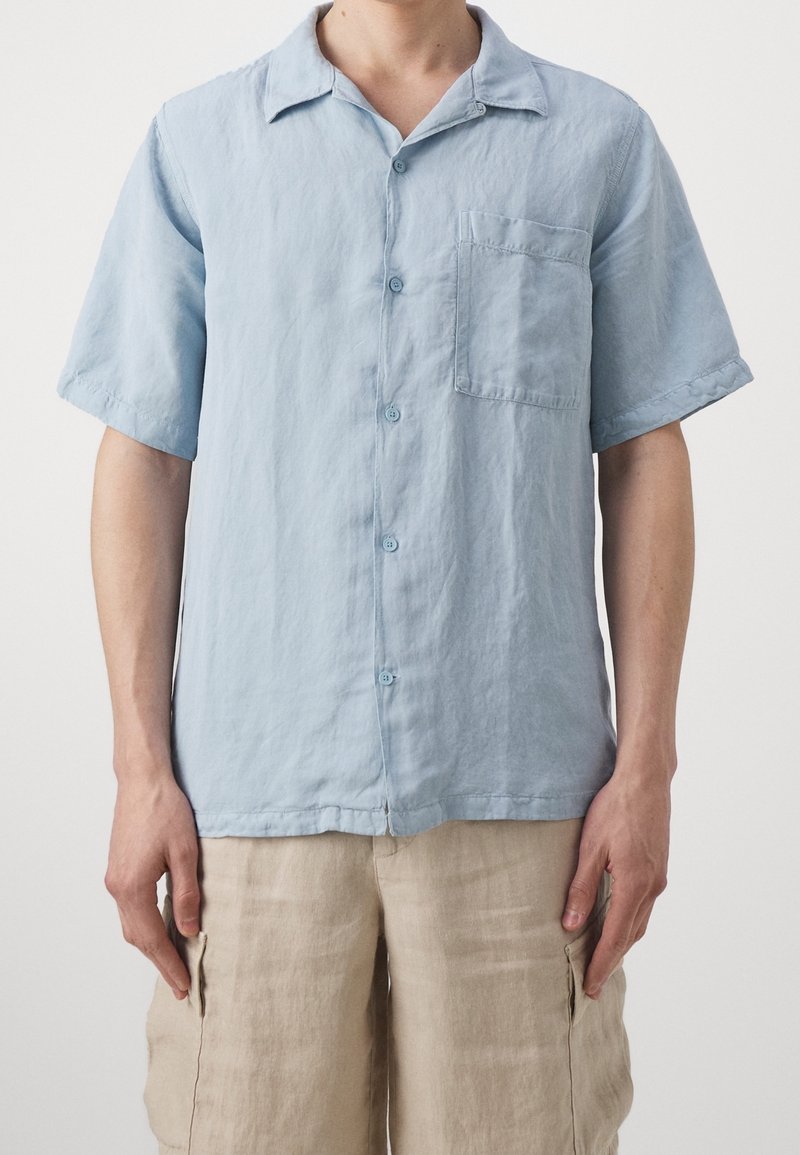 Camicia azzurra chiara a maniche corte in un tessuto texturizzato, con chiusura a bottoni, colletto e taschino sul petto. Indossata con pantaloni corti beige.
