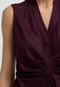 Blouse sans manches bordeaux avec un col en V, fermeture à boutons et détails de tissu froncé. Texture lisse avec une coupe ajustée.