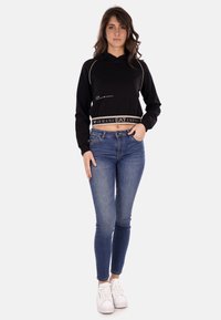 Felpa cropped nera con accenti beige e logo, abbinata a jeans skinny blu e sneakers bianche. Tessuto morbido, vestibilità casual.