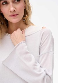 Hellgraues, langärmliges Strickoberteil mit lockerer Passform, das breite Ärmel und ausgeschnittene Schulterdetails aufweist. Die strukturiert wirkende Stoffoberfläche ist sichtbar.