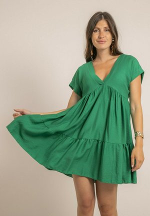 Femme aux longs cheveux bruns portant une robe courte et ample de couleur verte, tenant un côté de la robe pour montrer son tissu fluide.