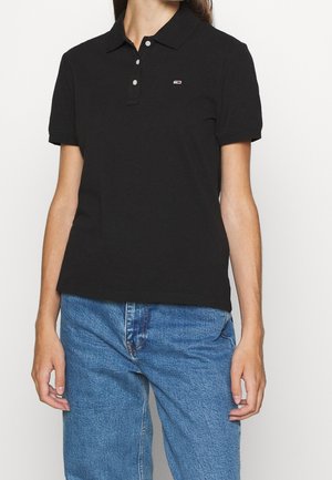 Woman wearing a black short-sleeve polo shirt with three buttons and blue denim jeans, shown from neck to hips.