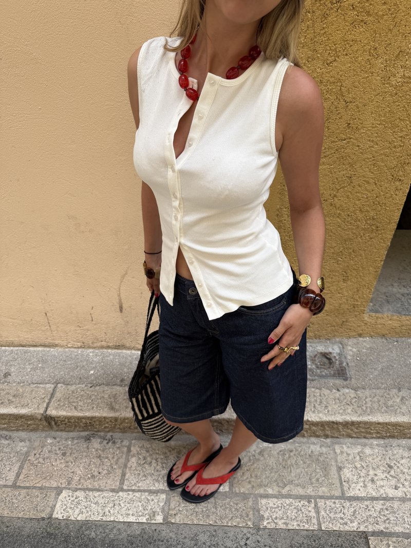 Femme portant un haut blanc sans manches à boutons, un short foncé au genou, un collier rouge et des sandales, debout sur un pavement en pierre près d'un mur jaune.