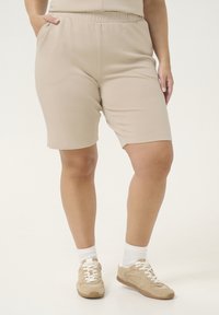 Shorts beiges à taille élastique avec poches latérales, tissu doux et texture lisse, associés à des baskets beiges et des chaussettes blanches.