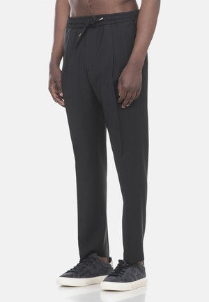 Uomo che indossa pantaloni neri con coulisse e sneakers in suede nero con suola bianca, in piedi davanti a uno sfondo chiaro e semplice.