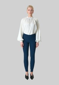 Witte blouse met lange mouwen en een geplooide halslijn, gecombineerd met strakke marineblauwe broek. Model draagt zwarte schoenen met puntige neus en gouden oorbellen.