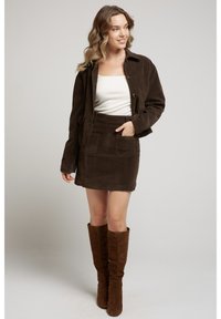 Femme portant une veste en daim brun foncé et une jupe avec des bottes hautes, haut crème, posant avec la main dans la poche de la jupe, souriante et regardant à droite.