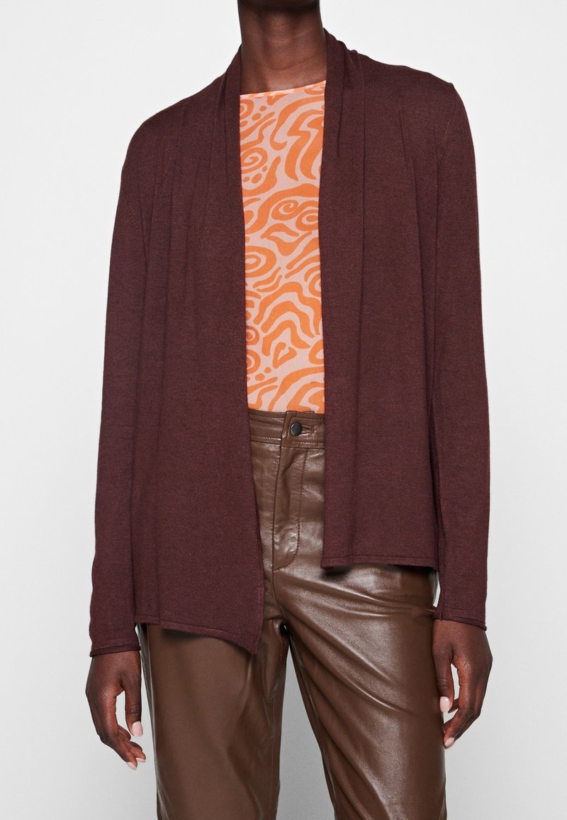 Personne portant un cardigan ouvert marron foncé, un haut orange à motifs et un pantalon en cuir marron, debout devant un fond clair uni.