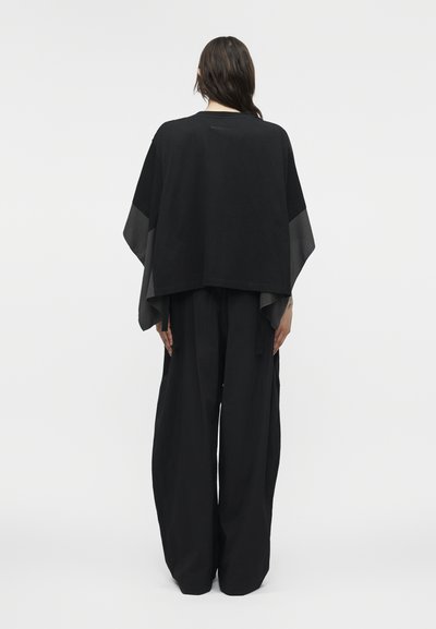 Άτομο με μακριά σκούρα μαλλιά φοράει φαρδιά μαύρη παντελόνα και χαλαρό μαύρο τοπ με φαρδιά μανίκια, στέκεται μπροστά σε απλό φόντο.