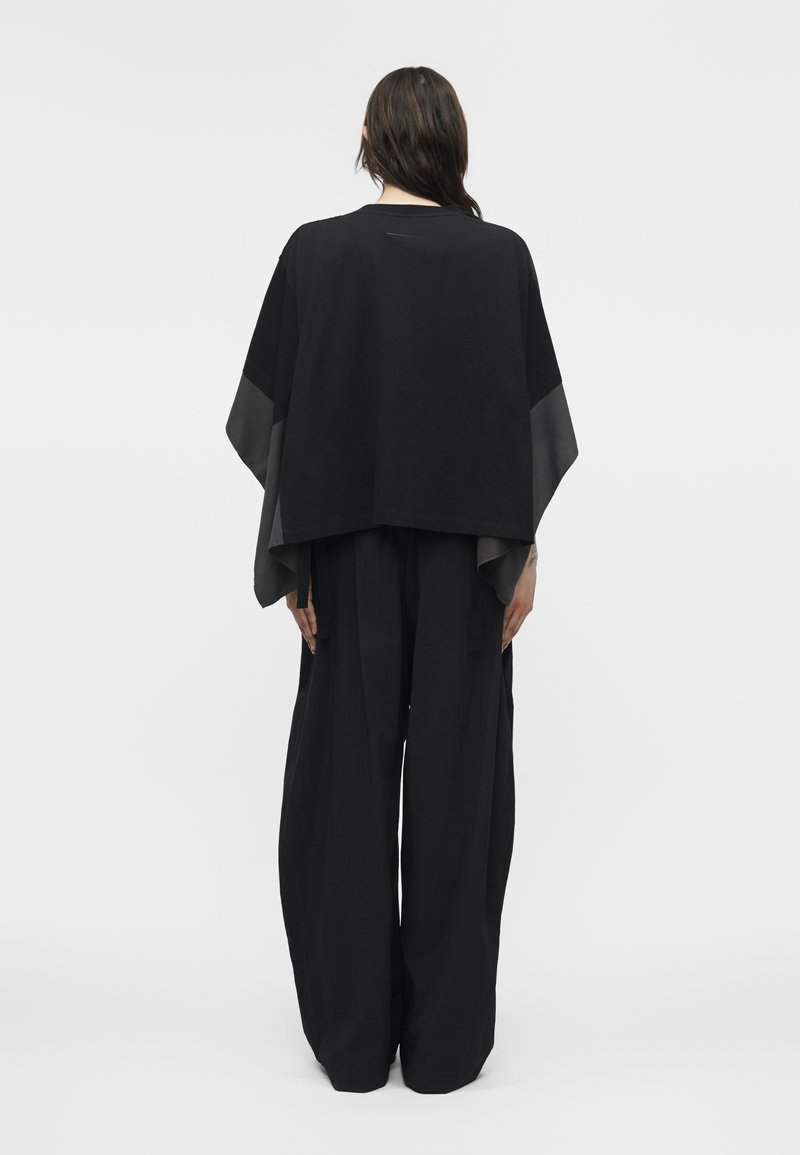 Personne aux longs cheveux noirs portant un pantalon large et ample noir et un haut noir drapé avec des manches larges, debout devant un fond uni.