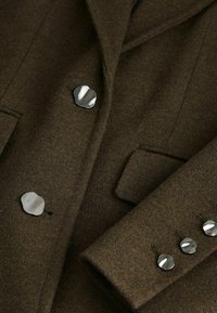 Hnědý vlněný blazer s klopami, který má stříbrné knoflíky na přední části a na manžetách, texturovanou tkaninu a boční kapsy.