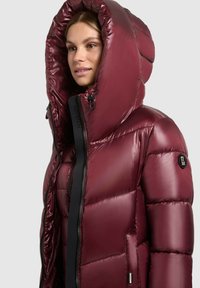 Chaqueta acolchada burdeos con una gran capucha, textura brillante, bolsillos laterales y detalle de cremallera negra; cuenta con un parche de logo en la manga.