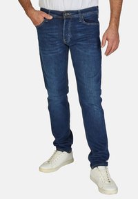Jeans de mezclilla azul oscuro con corte slim, estilo de cinco bolsillos, cremallera frontal y cierre clásico de botón. Llevados con zapatillas blancas.