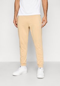 Világosbézs színű, lágy anyagból készült, sima tapintású, keskenyedő szabású melegítőnadrág. Rugalmas mandzsettákkal, oldalzsebekkel és karcsúsított fazonnal rendelkezik.