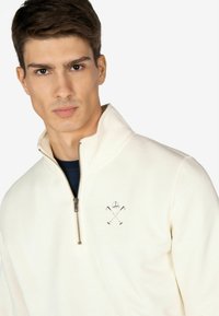 Maglione zip-up color crema con colletto alto, caratterizzato da un piccolo logo ricamato e una consistenza liscia. La cerniera presenta un accento metallico.
